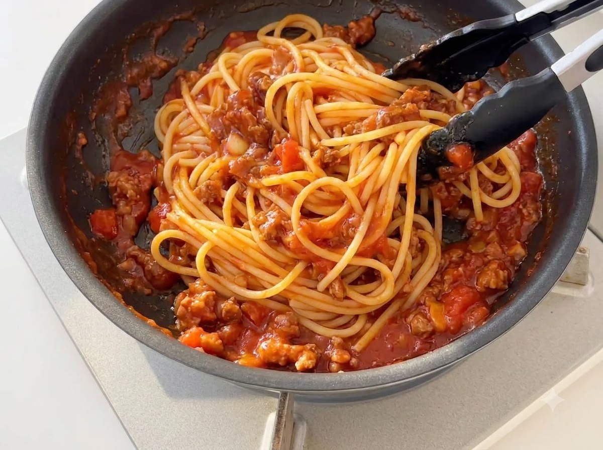 Spaghetti cotti mescolati con la salsa di pomodoro e carne addensata in una padella usando pinze in silicone nero.