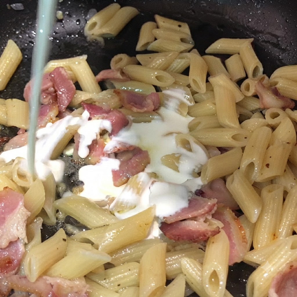 Latte e panna da montare versati dallalto in una padella riempita con pasta penne e pezzi di bacon.