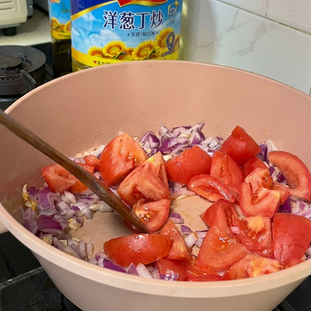 Pezzi di pomodoro fresco mescolati con cipolle rosse a cubetti e aglio in una padella rosa con una spatola di legno.