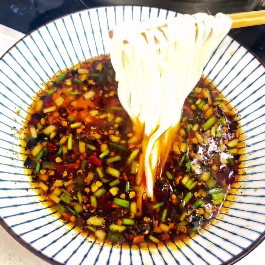 Delle bacchette sollevano una porzione di noodles bianchi cotti da una ciotola piena di brodo.