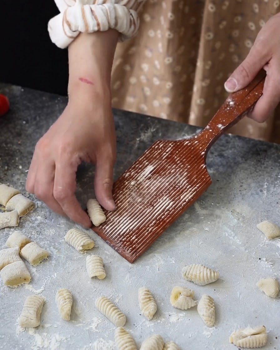Una mano che fa rotolare un piccolo pezzetto di pasta su un riga-gnocchi di legno scanalato.