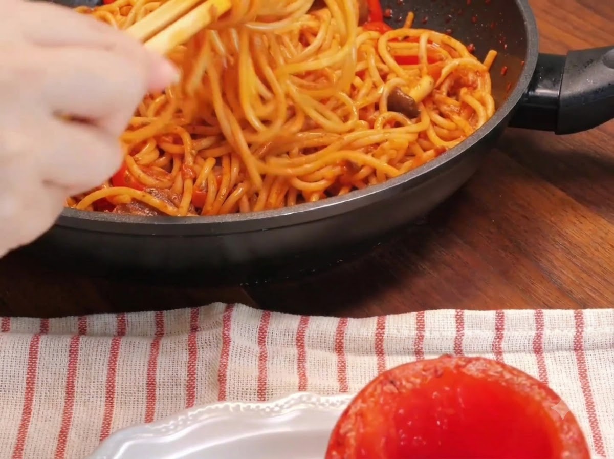 Un primo piano di una mano che usa le bacchette per arrotolare i noodles di spaghetti dentro una padella vicino a un pomodoro arrosto scavato su un piatto.