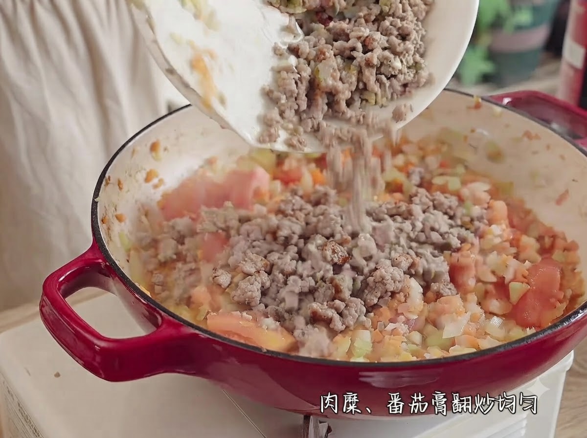 Carne macinata versata da un piatto bianco in una pentola rossa piena di pomodori, cipolle, sedano e carote a pezzi.