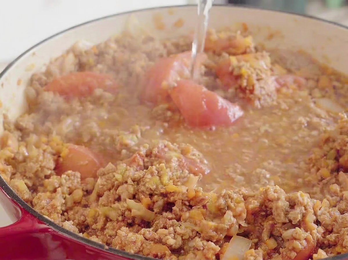 Acqua versata dallalto in una grande pentola rossa contenente il composto di carne, pomodoro e verdure in ebollizione.