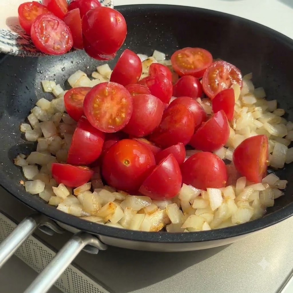 Pomodorini tagliati a metà aggiunti nella padella con cipolle e aglio saltati, mescolati insieme.