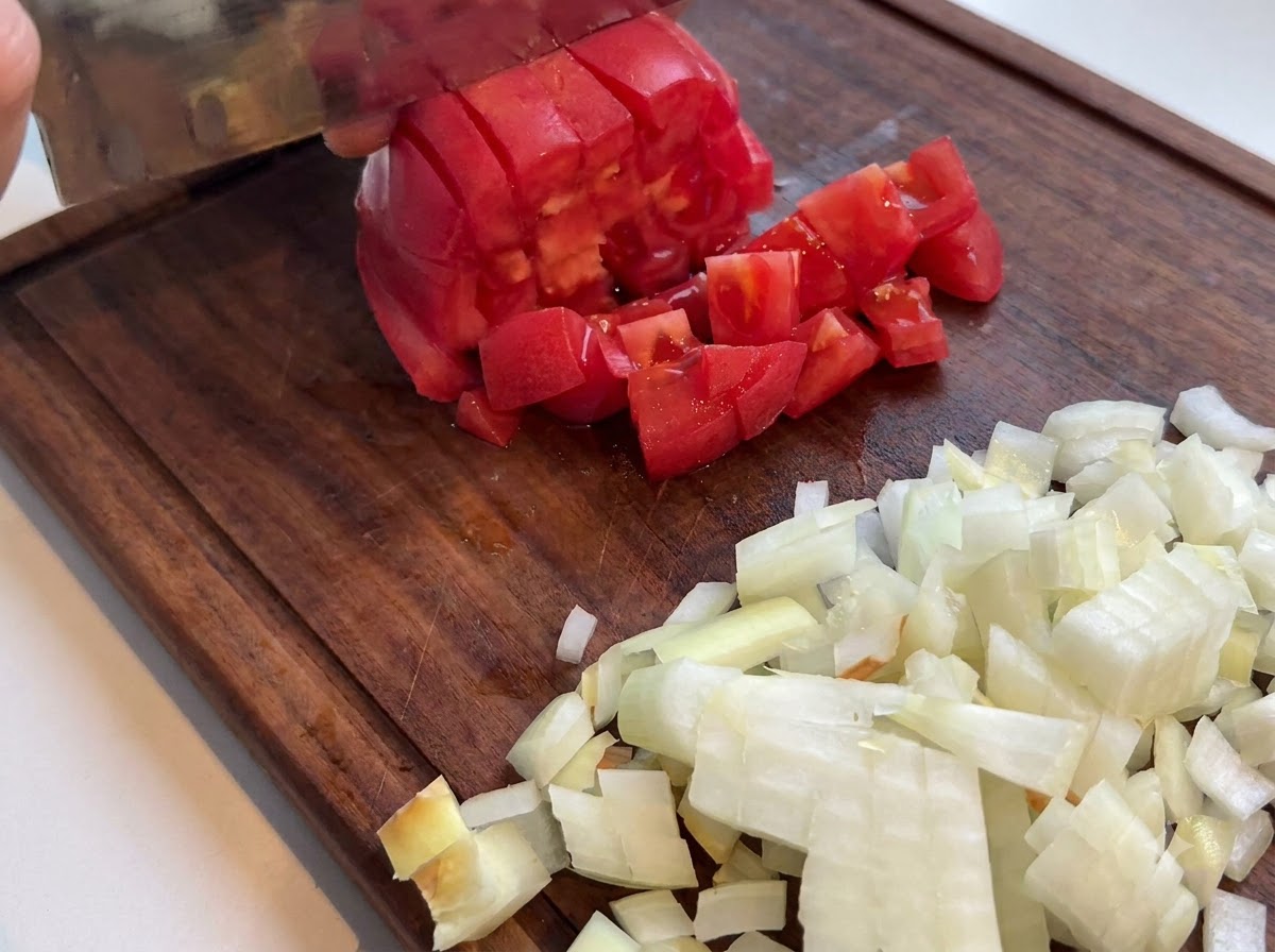 Pomodoro rosso fresco tagliato a cubetti con un coltello da cucina su un tagliere di legno vicino a cipolle tritate.