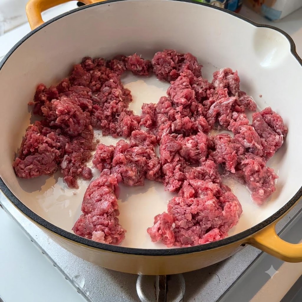Carne macinata di manzo cruda e cipolle bianche che sfrigolano in una padella dai toni chiari sul fuoco.