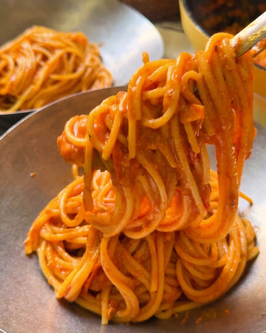 Pinze che sollevano una porzione di spaghetti cremosi al pomodoro in una ciotola di servizio in metallo lucido.