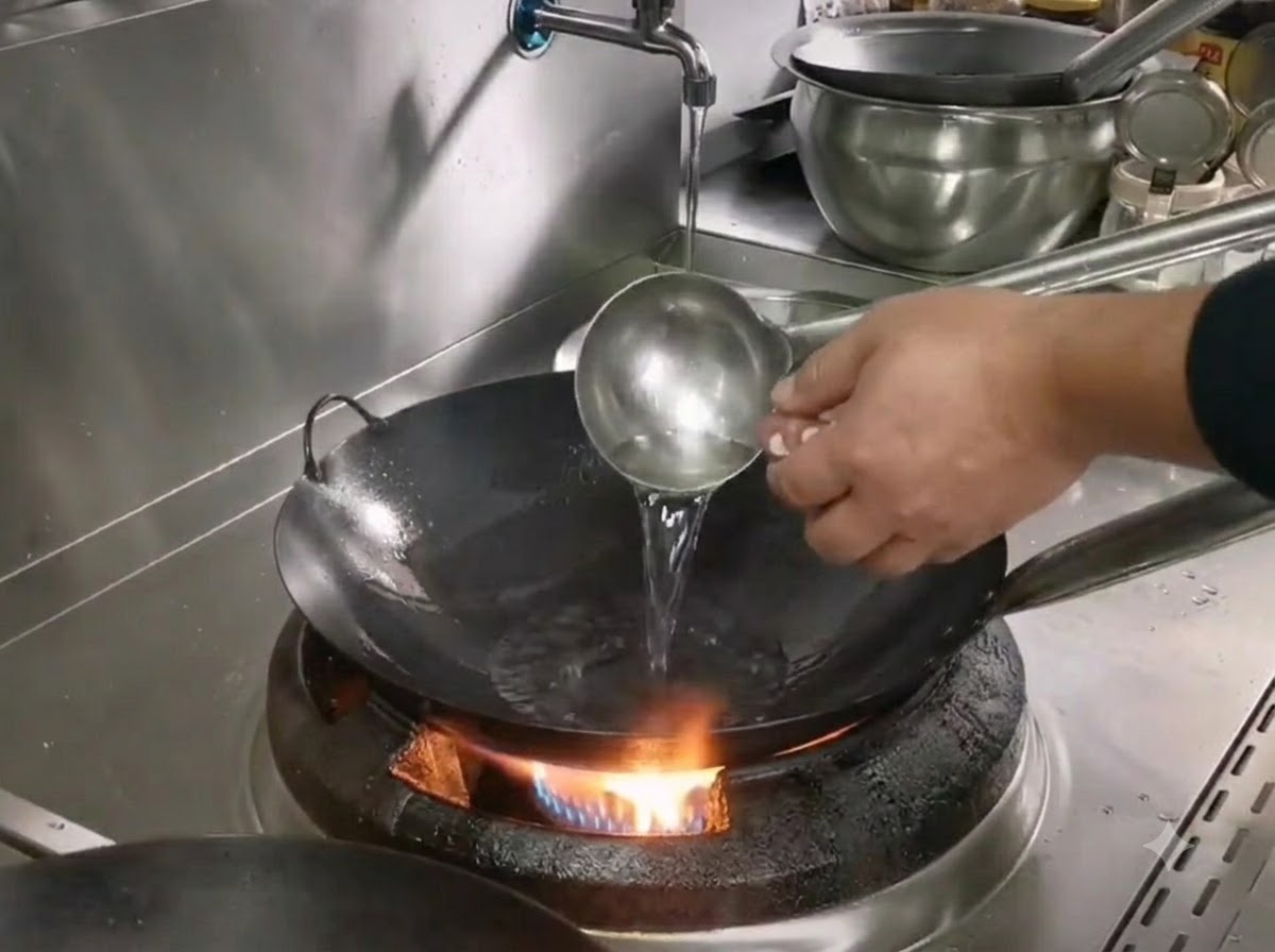 Un mettolino che versa olio in un wok di ferro caldo sopra un fornello a gas mentre si prepara a cuocere il maiale sminuzzato.