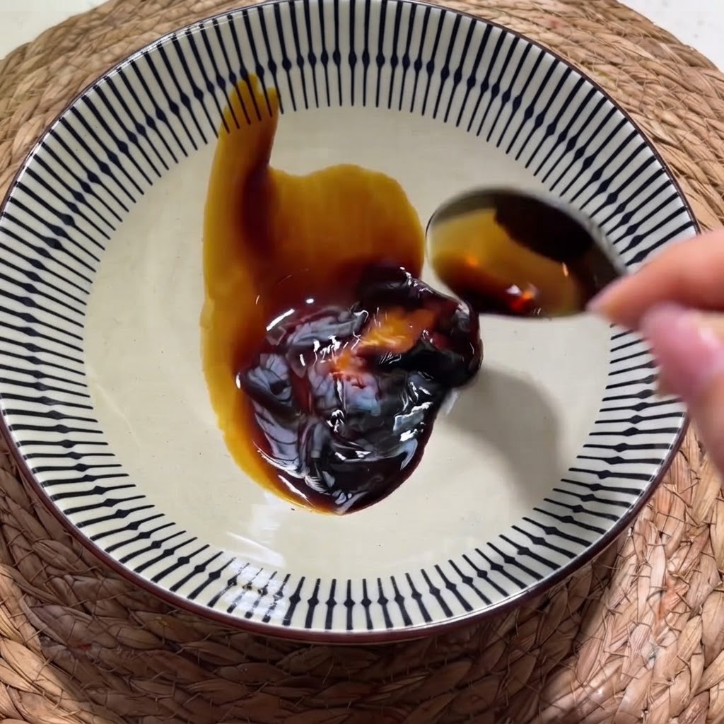 Una mano che versa salsa di soia scura da un cucchiaio in una ciotola di ceramica decorata contenente una noce di salsa di ostriche.
