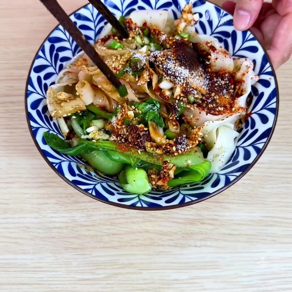 Bacchette che mescolano i noodles larghi di grano, il bok choy e la salsa di olio al peperoncino insieme allinterno della ciotola di ceramica.