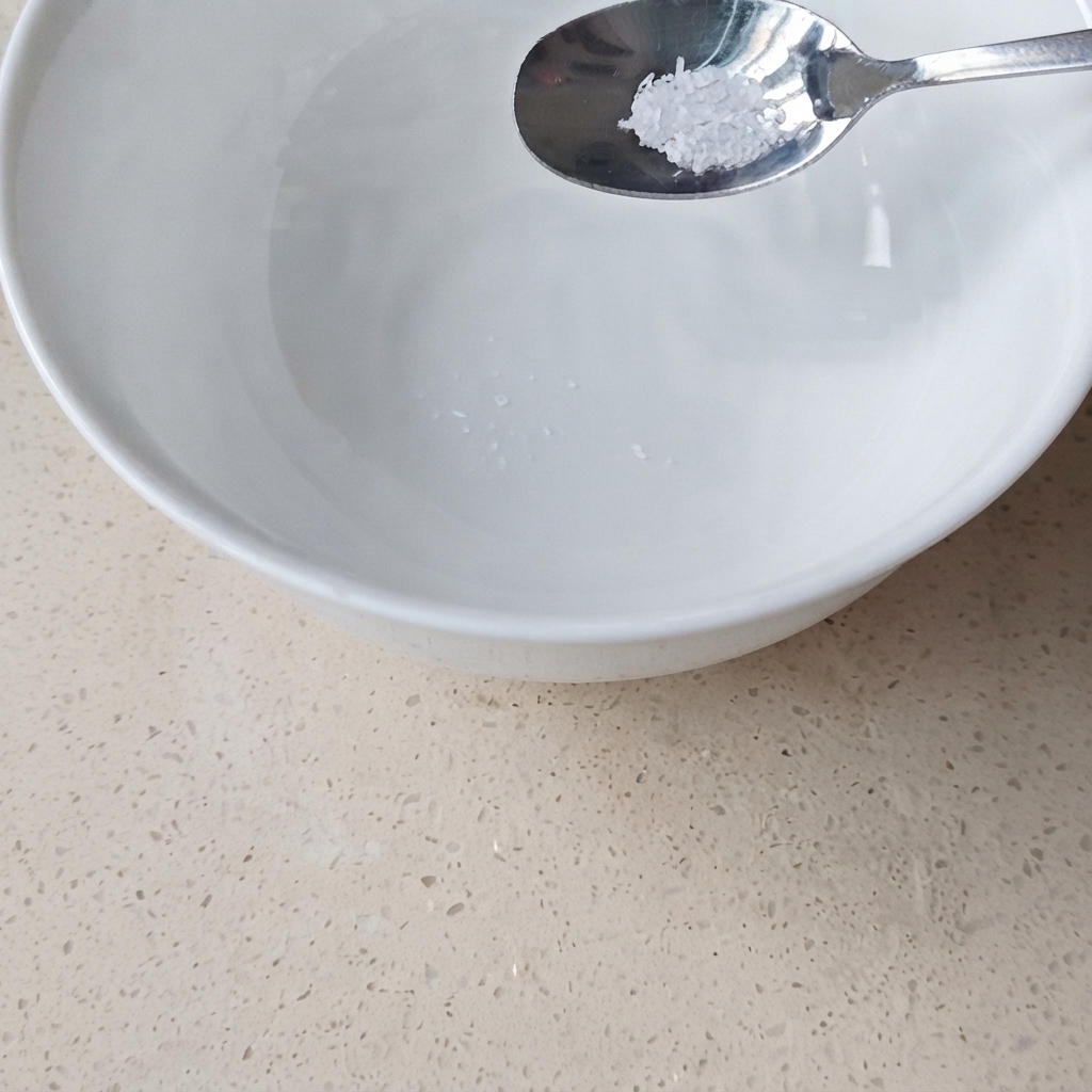 Un cucchiaio di metallo con una piccola quantità di glutammato monosodico posizionato sopra una ciotola di ceramica bianca contenente acqua.