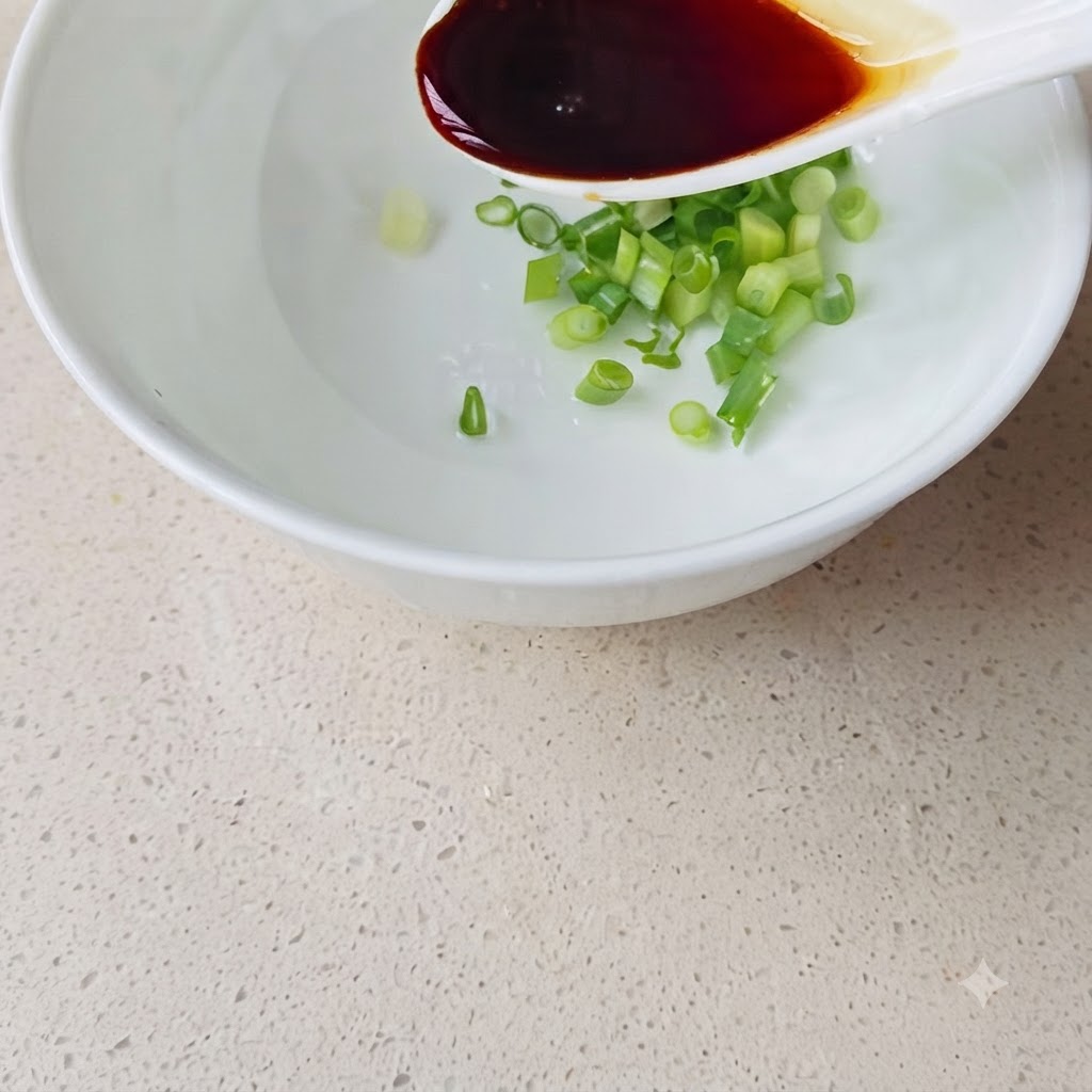 Un cucchiaio bianco versa della salsa di soia scura in una ciotola bianca contenente cipollotto tritato e acqua.
