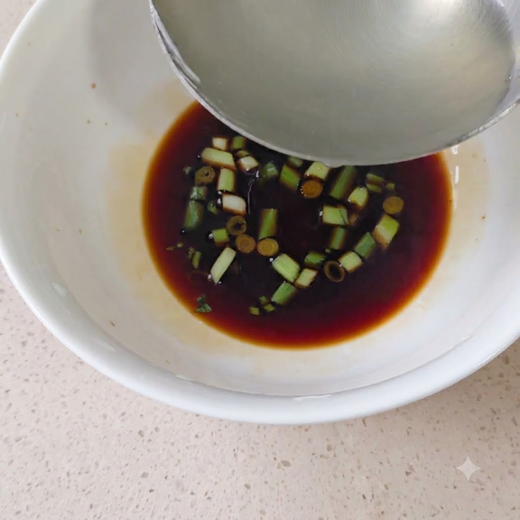Un mestolo di metallo versa del brodo caldo in una ciotola bianca piena di salsa di soia e cipollotto tritato.