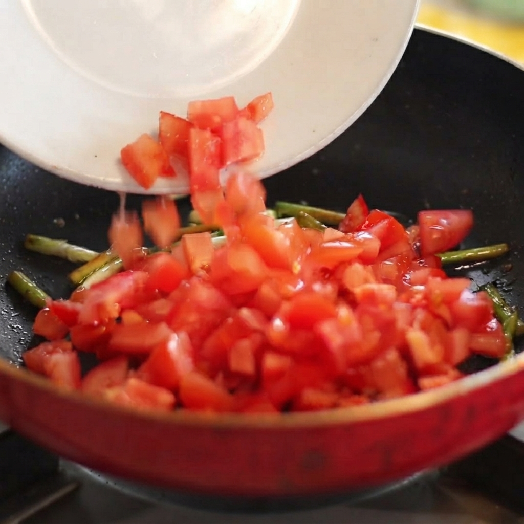 Pomodori a dadini che scivolano da un piatto bianco in una padella calda con cipollotti saltati.