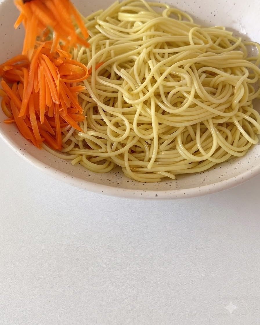 Carote fresche sfilacciate e altri condimenti disposti sopra i noodles cotti in una grande ciotola di ceramica.