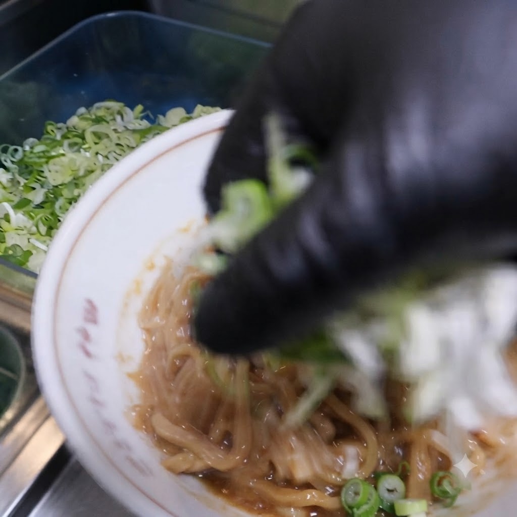 Una mano guantata che sparge cipollotti ed erba cipollina tritati sopra i noodles conditi nella ciotola.