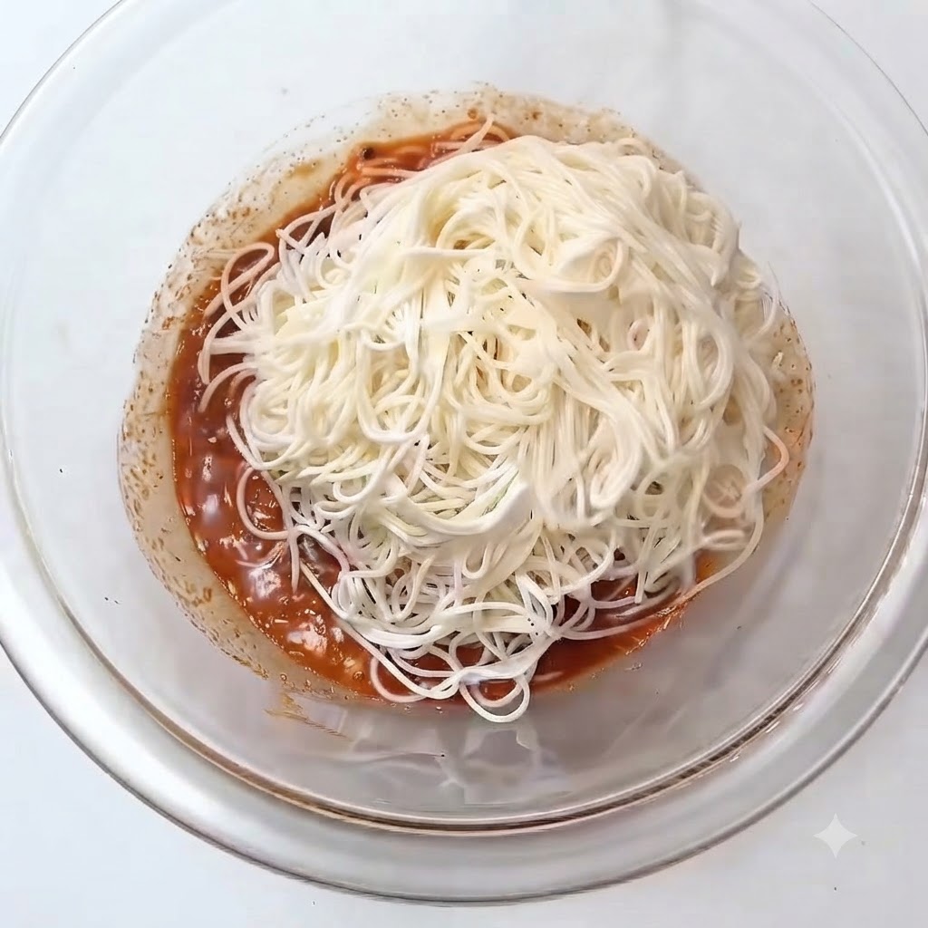Una mucchietto di spaghetti somen bianchi cotti posti sopra la salsa rossastra allinterno di una ciotola di vetro.
