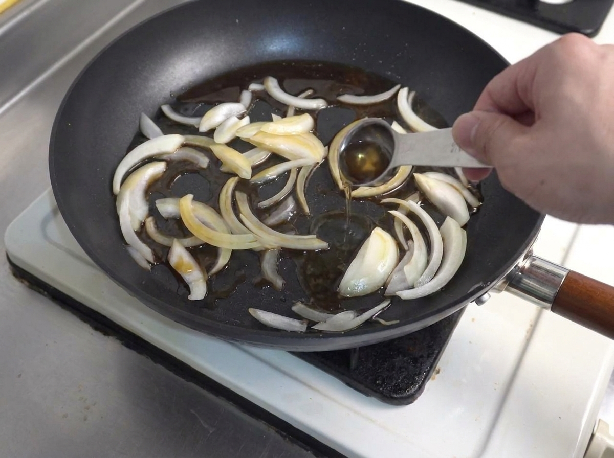 Un cucchiaio misuratore che versa condimento liquido in una padella con fette di cipolla saltate.