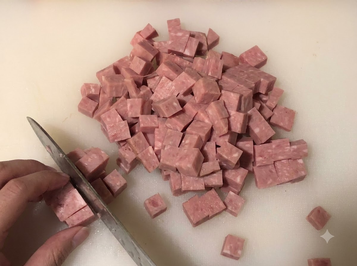 Spam tagliato a piccoli cubetti su un tagliere con cipolle tritate e cipollotti sullo sfondo.