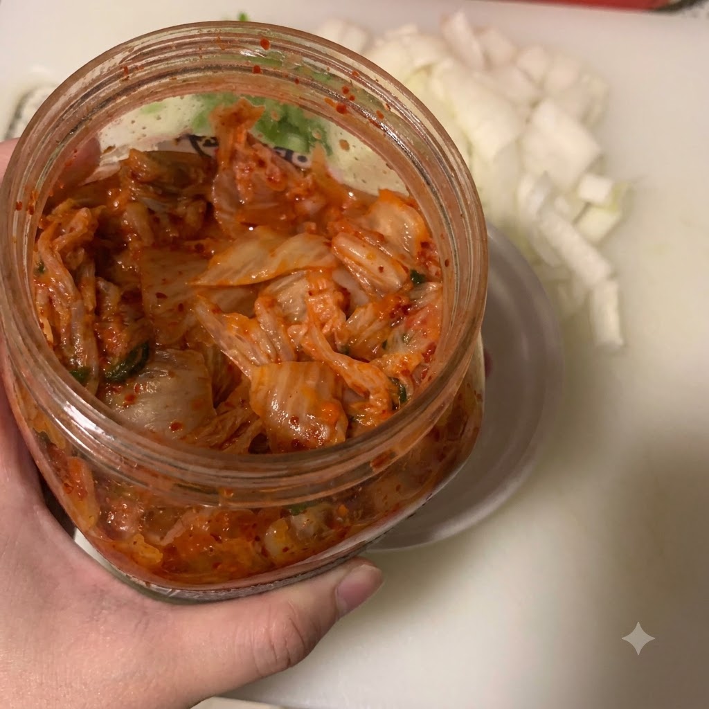 Kimchi trasferito in una ciotolina bianca con un cucchiaio accanto a un contenitore di cipolle tritate.