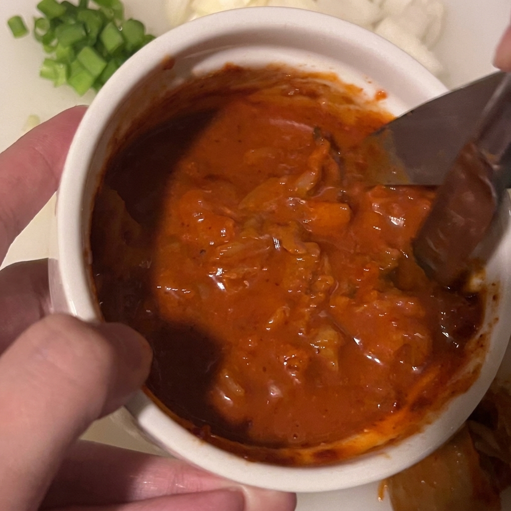 Forbici da cucina che tagliano pezzi di kimchi sommersi in un denso sugo rosso denso in una ciotolina bianca.