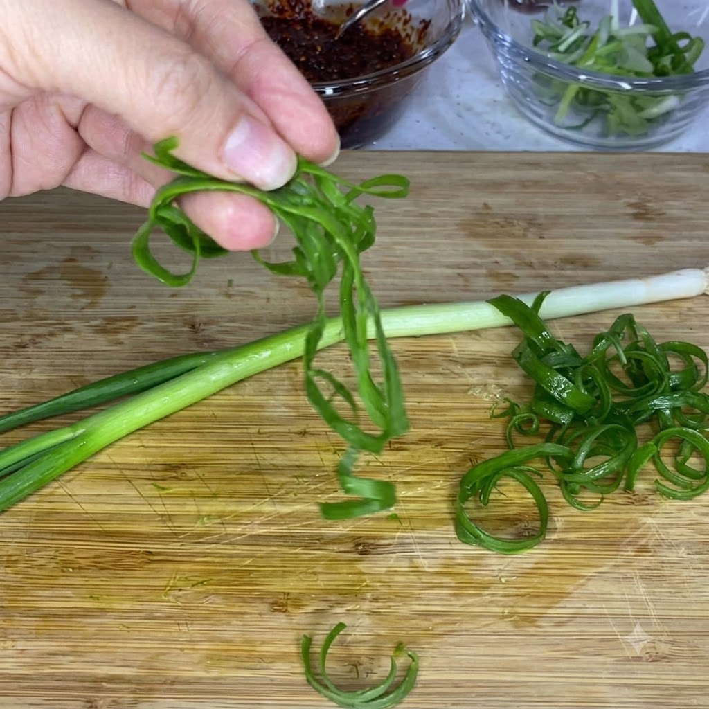 Una mano che tiene strisce di cipollotto verde arricciate sopra un tagliere di legno, con ciotole di vetro di salsa e cipolle affettate sullo sfondo.