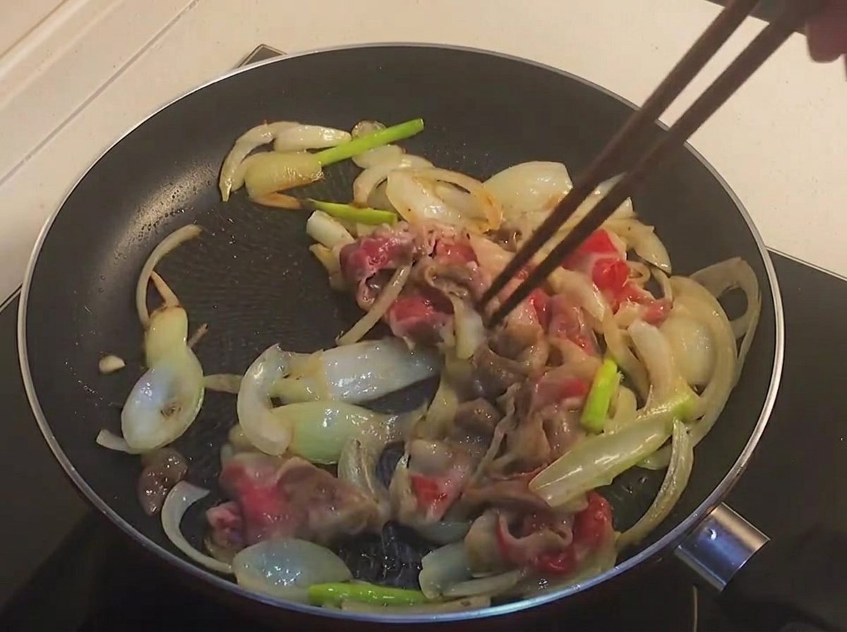 Bacchette che mescolano fettine di pancetta di manzo parzialmente cotte e leggermente dorate con le cipolle in una padella nera.