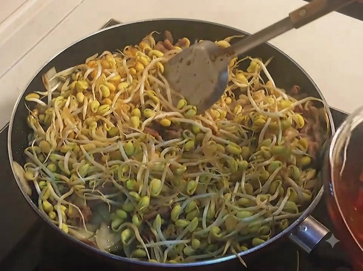 Un mestolo che versa una salsa rossa scura piccante su una padella piena di germogli di soia gialli.