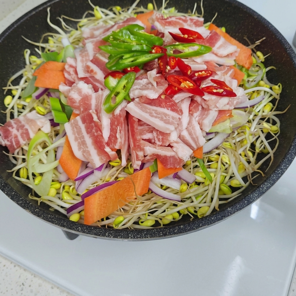 Una vista dallalto di una padella riempita con uno strato di germogli di soia sormontato da cipolle affettate, carote, strisce di pancetta di maiale cruda e fette di peperoncino verde e rosso.