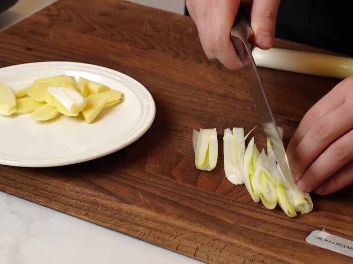 Uno chef che affetta cipollotti e pezzi di zenzero su un tagliere di legno accanto a un piatto bianco.