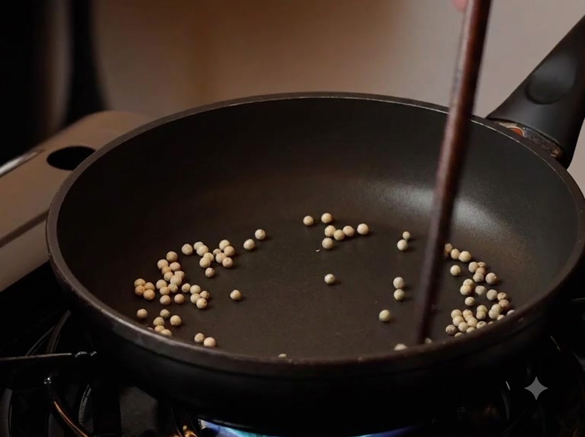 Grani di pepe bianco tostati in una padella nera con le bacchette che li mescolano a fuoco basso.