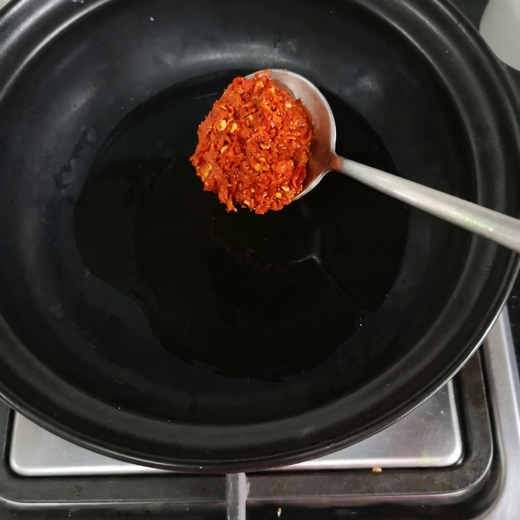 Un cucchiaio di pasta di peperoncino rosso inserito in una pentola di terracotta nera sul fuoco.
