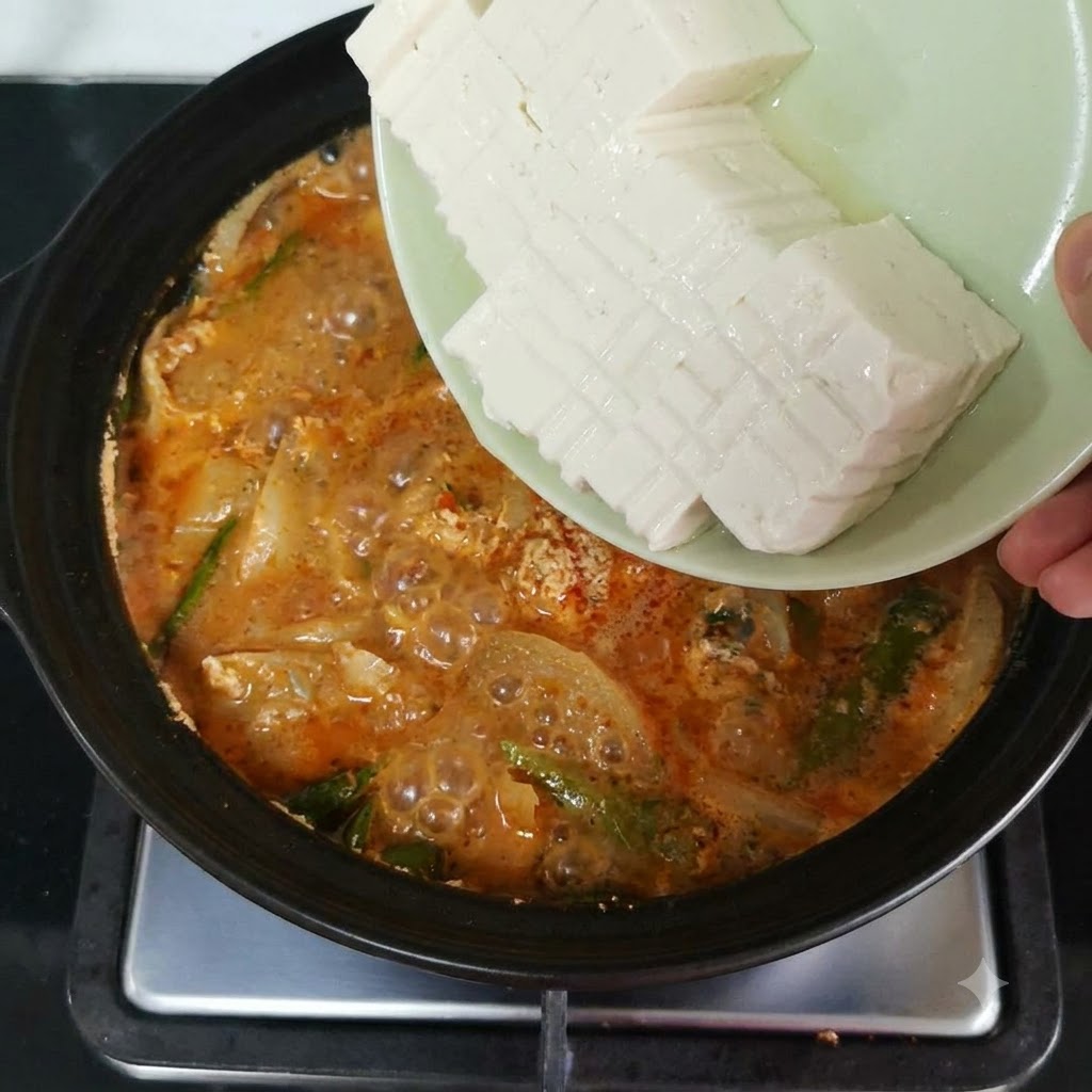 Fette di tofu morbido fatte scivolare da un piatto in una pentola in ebollizione piena di brodo rosso-arancio e verdure.