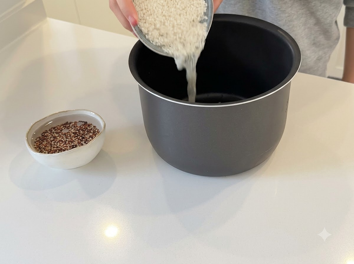 Mani che versano riso bianco nella pentola scura di un cuociriso con una piccola ciotola di quinoa in ammollo vicino.