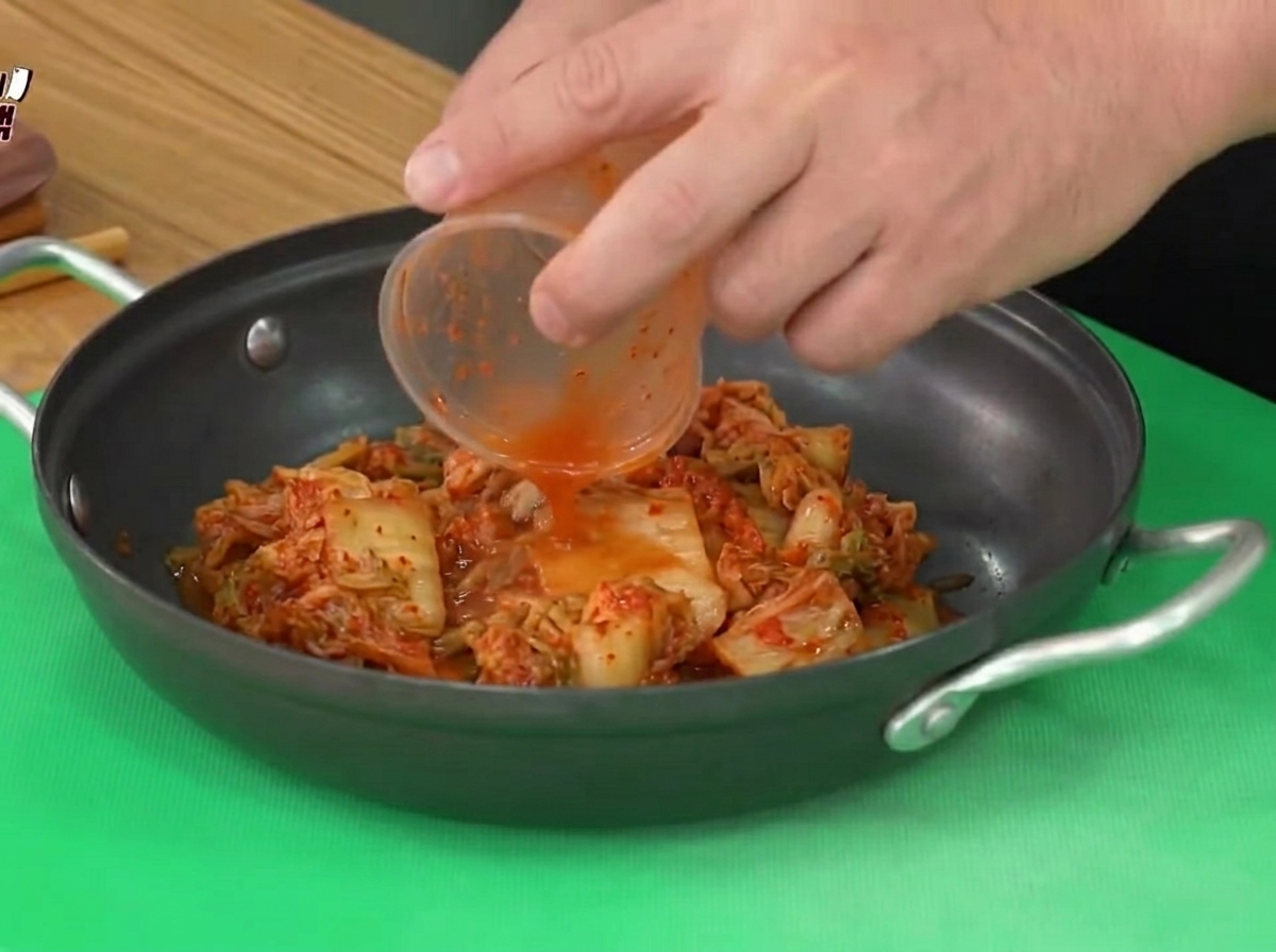 Kimchi aspro aggiunto da un contenitore di plastica in una pentola da cucina nera.