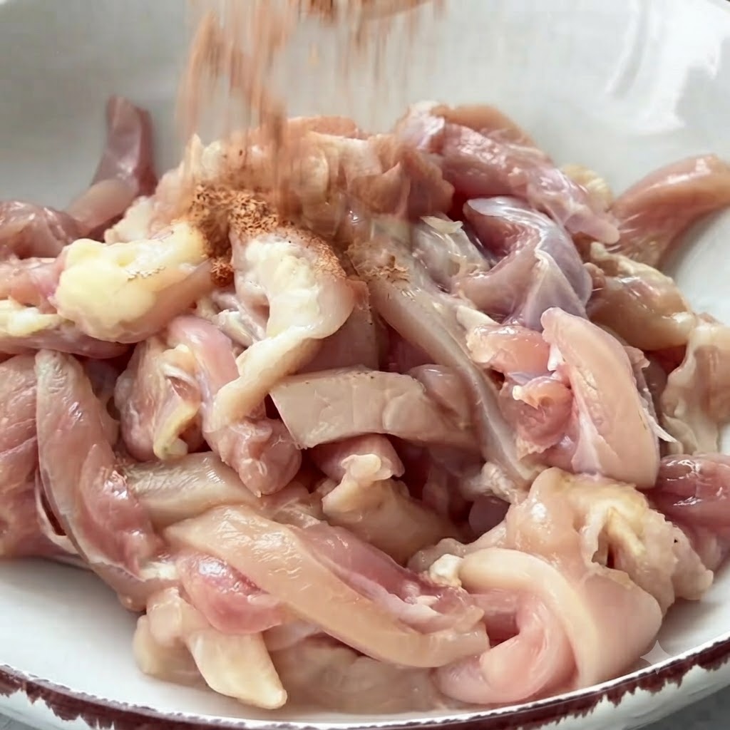 Spezie versate in una ciotola piena di straccetti di pollo crudi pronti per essere mescolati.