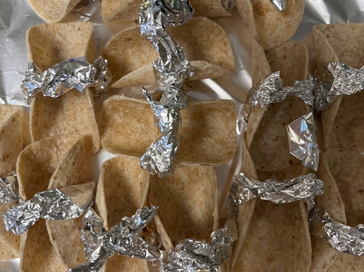 Piccoli involucri di tortilla disposti e fissati con pezzi di alluminio su una teglia pronti per il forno.