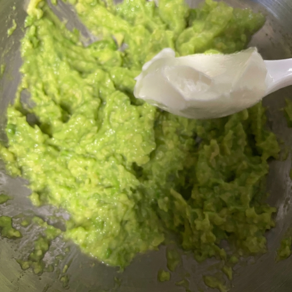 Un cucchiaio bianco che raccoglie yogurt greco denso e lo aggiunge in una ciotola di avocado appena schiacciato.
