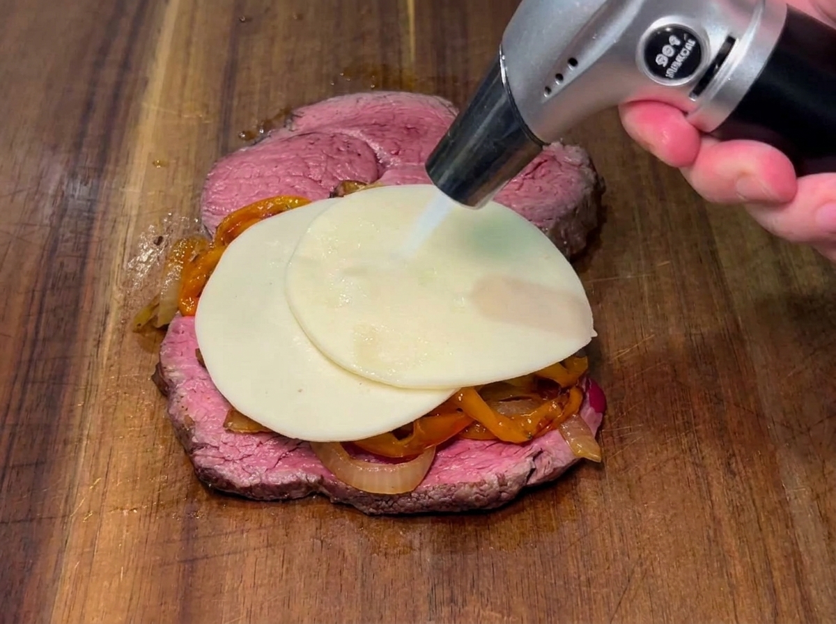 Una fetta di formaggio fatta sciogliere con un cannello a gas sopra i peperoni e le cipolle su una bistecca.