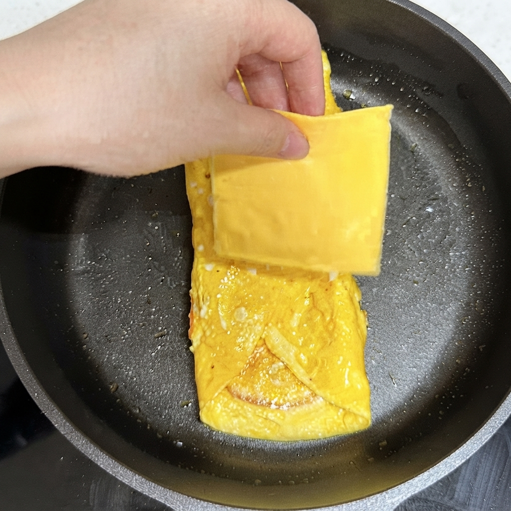 Una mano che posiziona una fetta quadrata di formaggio giallo sul pacchetto di uova cotto nella padella.