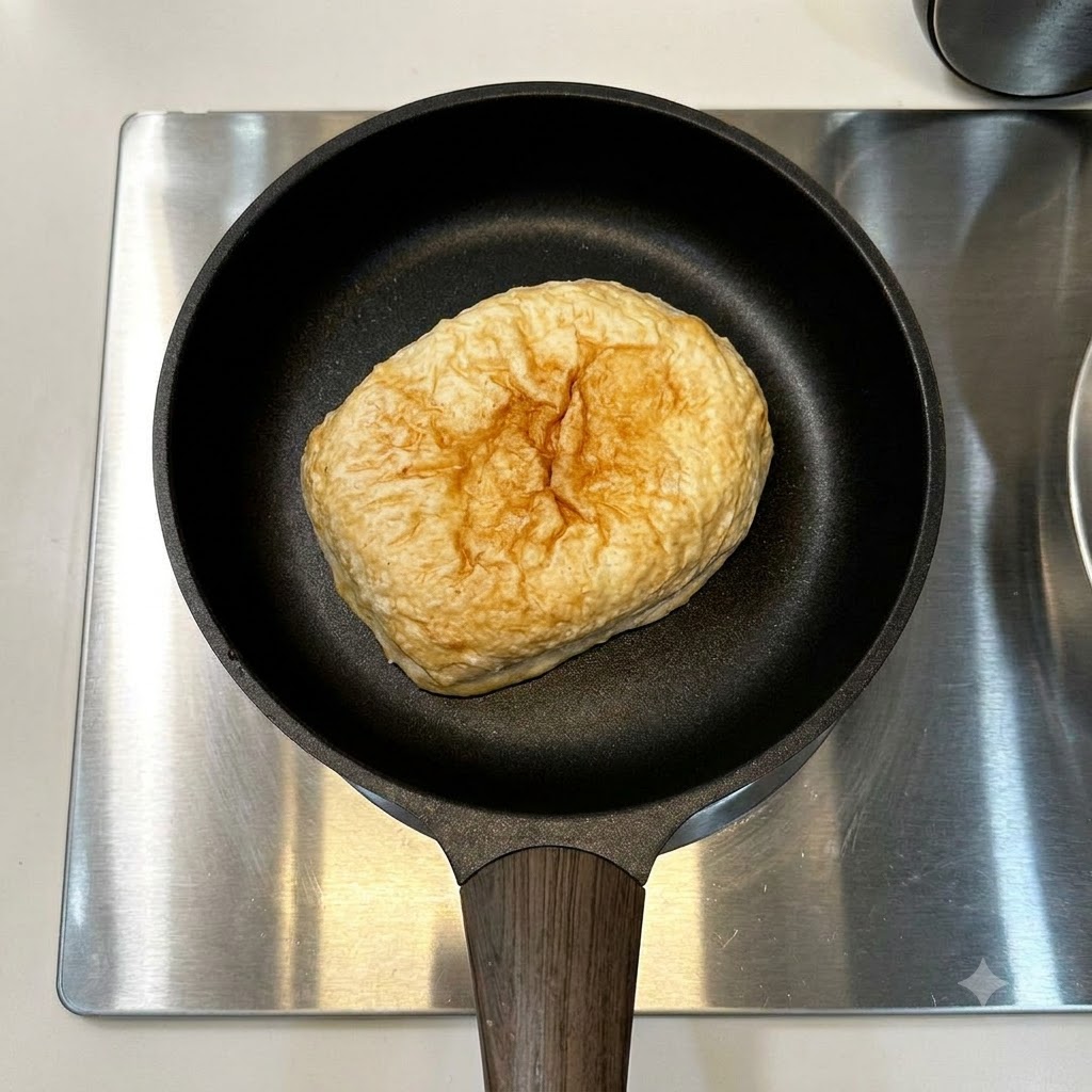 Pane ciabatta e una singola salsiccia adagiate in una padella nera su un piano cottura in acciaio inossidabile.