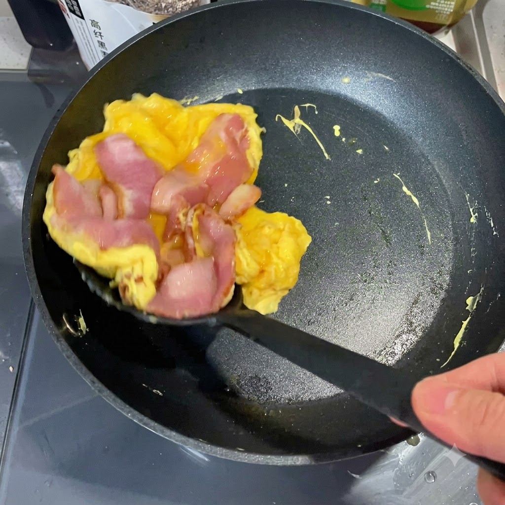 Una spatola che solleva una pila di uovo cotto e pancetta da una padella per posizionarla su una metà di muffin inglese tostato.