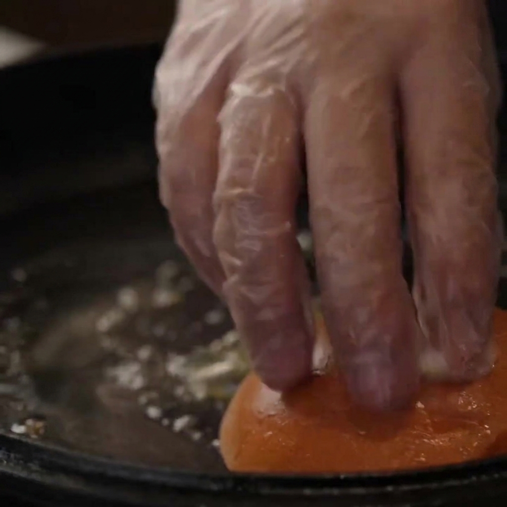 Una mano guantata che preme i panini per hamburger verso il basso in una padella di ghisa imburrata per toastarli.
