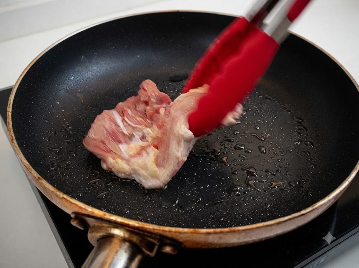 Una coscia di pollo cruda che viene inserita in una padella riscaldata con olio usando delle pinze rosse.