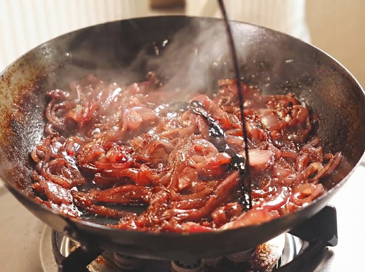 Cipolle rosse affettate che sfrigolano in un wok scuro con vino scuro e aceto versati sopra.