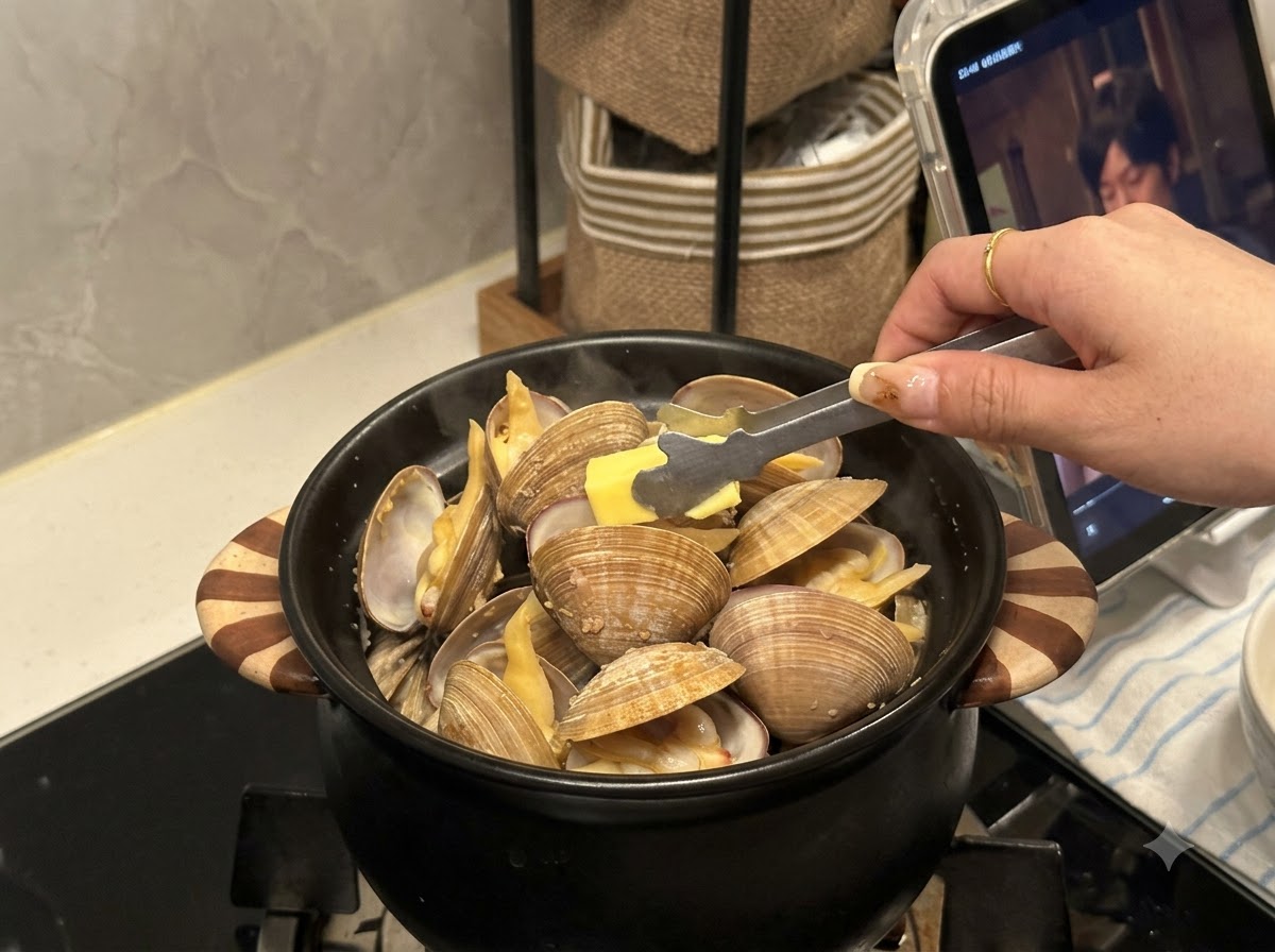 Un pezzetto di burro aggiunto con una pinza in un tegame nero pieno di vongole aperte.