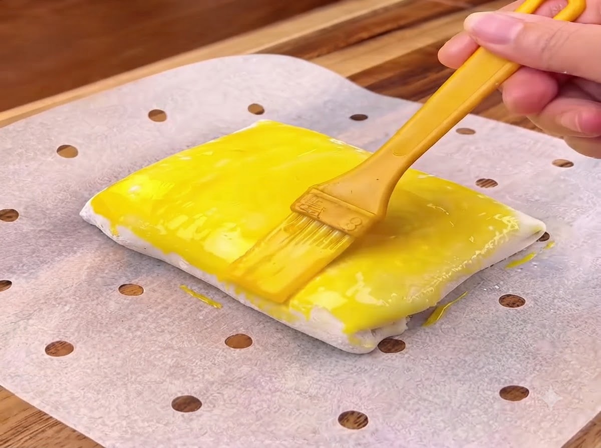 Un pennello giallo che applica luovo sbattuto sulla superficie di un quadrato di pasta sfoglia piegata.