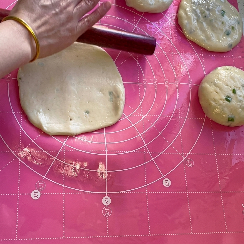 Un mattarello di legno che appiattisce una porzione di impasto in un cerchio sottile su un tappetino rosa.