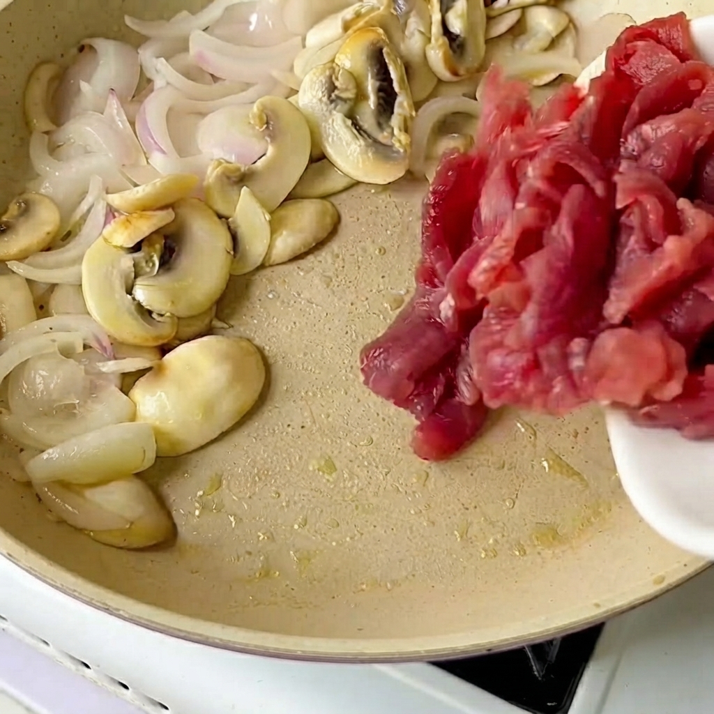 Manzo affettato trasferito da un piatto bianco in una padella con cipolle e funghi saltati.